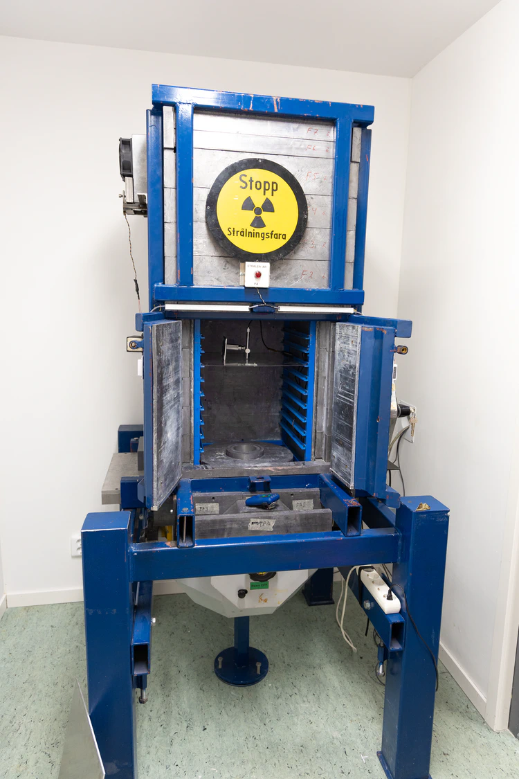 A large setup held together by blue steel profiles. There is a chamber in the middle with doors ajar, lead lined walls. Stacks of lead blocks are visible on the top of the chamber with a radiation warning sign in front.