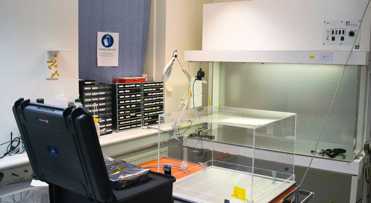 A bench with a stainless steel top. In front of the bench is a perforated metallic sheet. Above the bench are some lights lighting up the perforated sheet and the bench. On the left of the bench is a large magnifying glass mounted on a movable holder. In the foreground a black space hardware transportation box and a clear acrylic box.