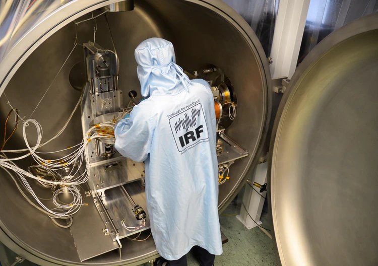 An open stainless steel chamber in the background revealing instrumentation along with wiring and a movable platform. A scientist in a blue IRF cleanroom coat in the foreground handling the instrumentation.