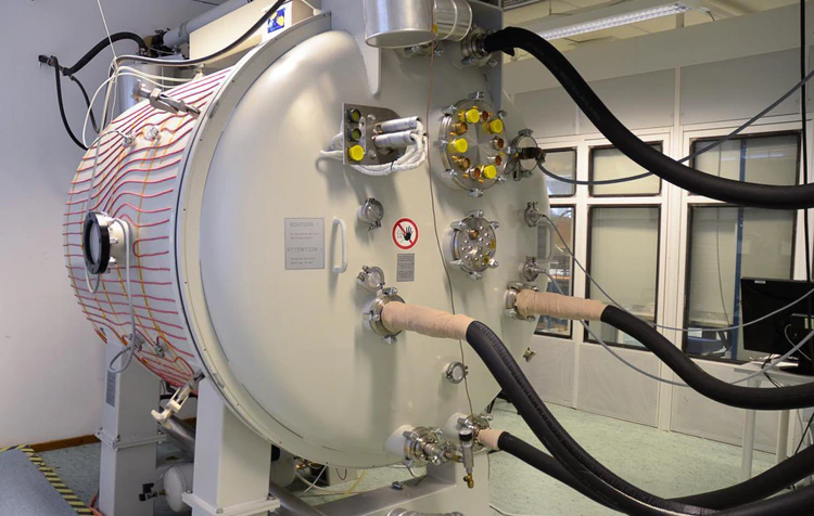 White cylindrical chamber oriented with its axis parallel to the ground. Several vacuum flanges visible on the door. Four insulated pipes carrying liquid nitrogen to the chamber visible in the foreground. A viewport is visible on the the circumference.