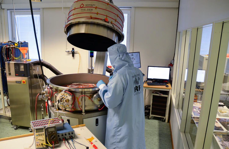Stainless steel chamber in the open position with top half supported using an arm and bottom half mounted on a sturdy table. A copper plate and wiring visible inside the chamber, with many vacuum flanges along the circumference. A researcher wearing a cleanroom coat looking into the chamber. Computers in the background to control the system. Oil control system visible on the left.