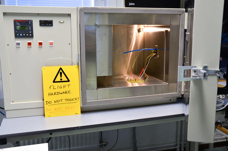 Two boxes attached to each other side-by-side. The white box on the left has the control panel with a display and operating buttons. In front of the box is a yellow warning sign. The metallic box on the right is open - the chamber inside with a PCB and other wires visible.