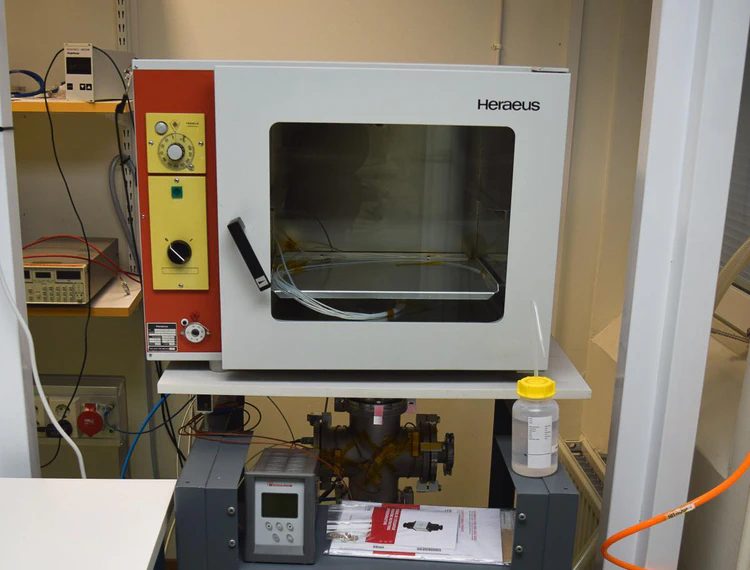 White baking oven with a glass viewport and door handle. On the left are the control knobs. The oven is mounted on a sturdy platform. Vacuum flanges are visible on the bottom.
