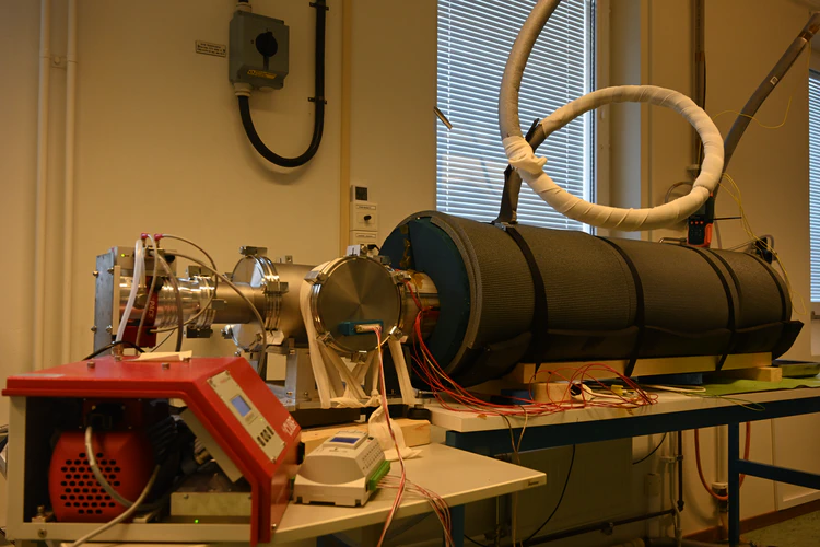 Stainless steel tube with multiple feed through flanges on the left. The pipe continues on the right but is covered with thick black insulation. Liquid nitrogen inlet and outlet pipes are visible on the top. A white data logger and a red vacuum pump are visible in the foreground.