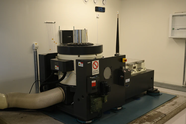 Large shaker in a room. It is currently in the Z direction. A test article is placed on the armature. A large pipe carrying air is visible on the bottom left.