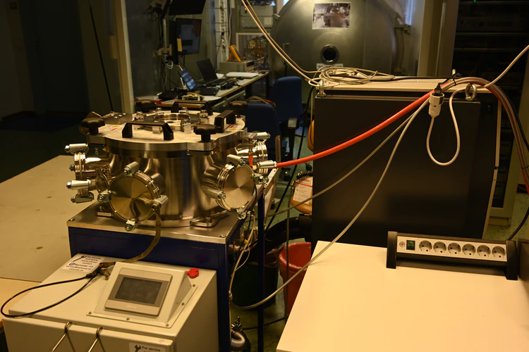 Stainless steel cylinder with six feedthrough flanges dstributed across its circumference (four visible) mounted on a table. A white control module visible in foreground while a black control module is visible on the right.